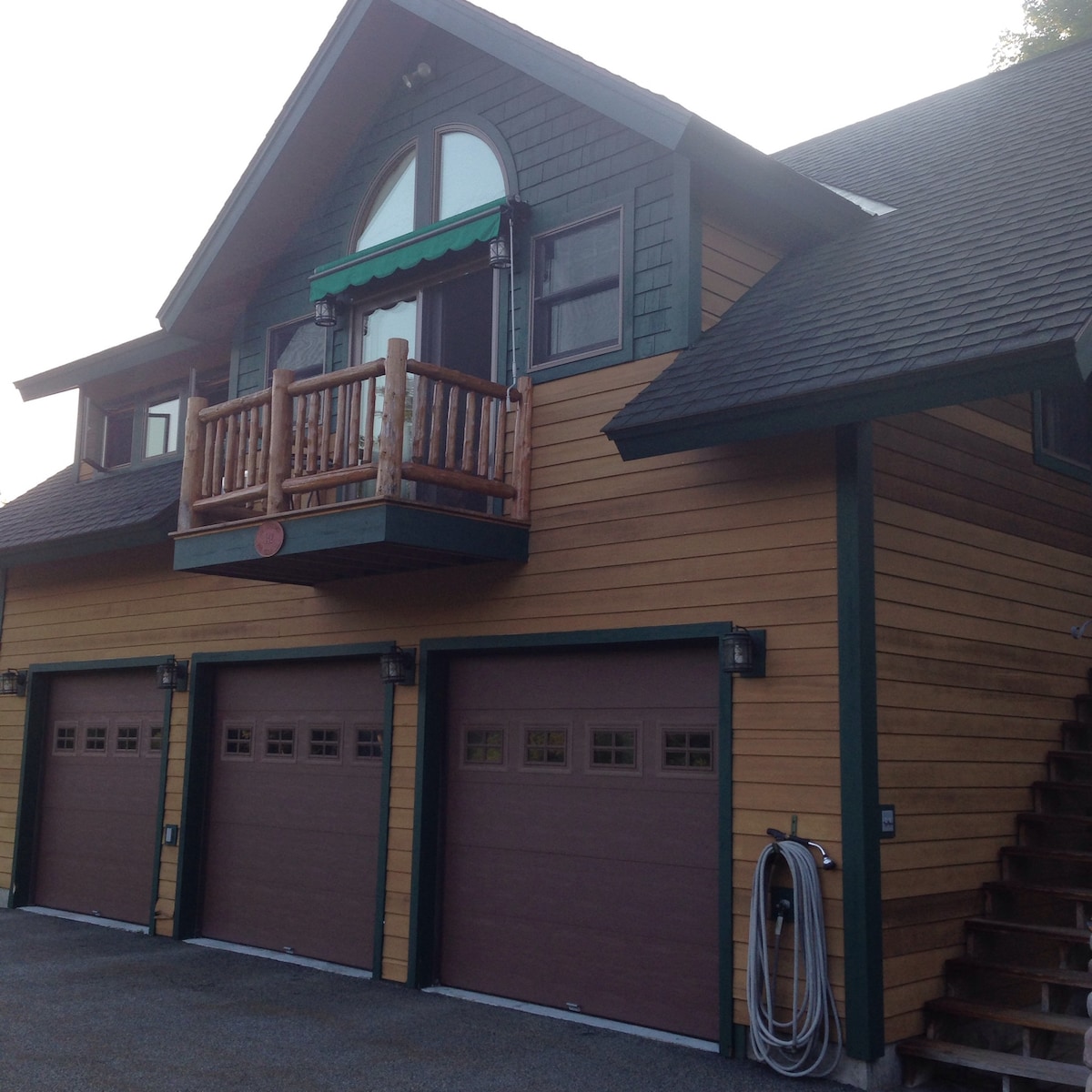 The exterior of the building is characterized by a combination of natural wood siding and dark green accents. A balcony is visible above three garage doors, which feature glass paneling. A staircase leads to the entrance beside the garage area.