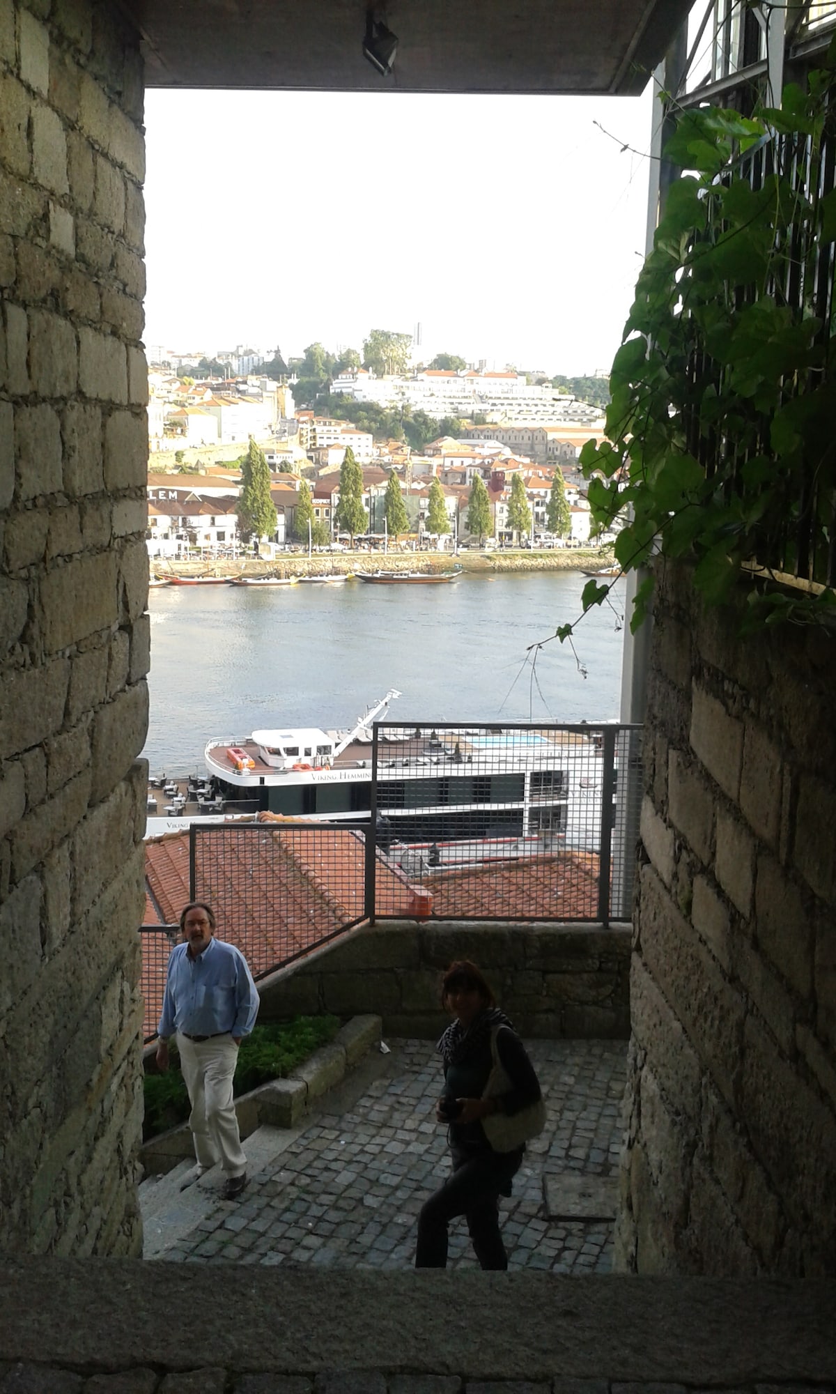 A stone staircase leads down to a riverside view, framed by robust stone walls. The water reflects the surroundings, with boats moored nearby. Vegetation climbs along the upper edges, adding a touch of greenery to the picturesque setting. The pathway is lined with cobblestones.