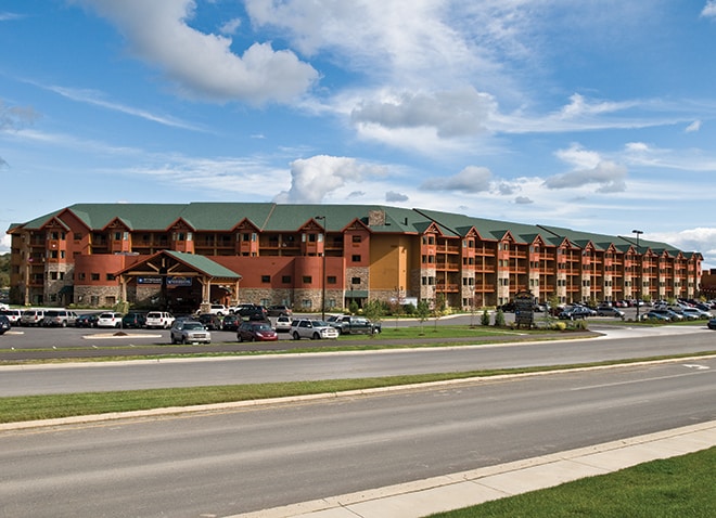 A large resort building features a blend of wood and stone finishes under a green roof. Multiple levels of balconies provide access to outdoor views. A spacious parking area is visible in front, surrounded by landscaped greenery and cars.