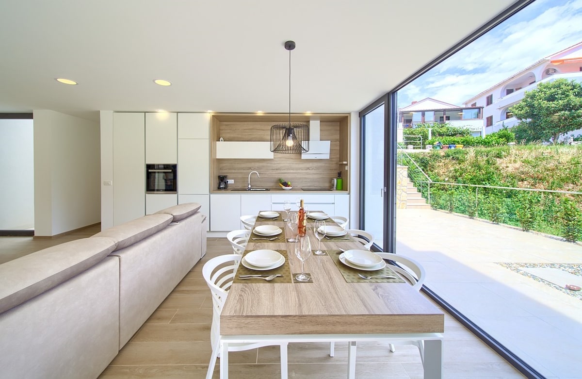 A modern dining area is presented with a wooden table set for six, featuring stylish tableware. The kitchen is visible in the background, equipped with sleek cabinetry and appliances. Large glass doors invite natural light and offer views of the garden outside.