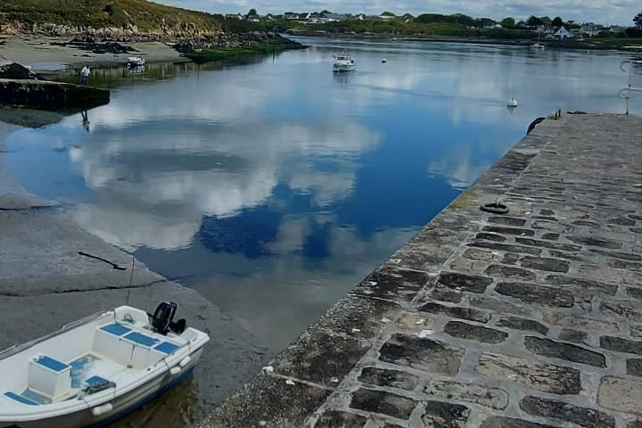 A serene view of a calm waterway is captured, reflecting soft clouds in the sky. A small boat is positioned along the stone pier, while other boats are visible in the distance. The landscape features lush greenery along the shoreline, enhancing the peaceful ambiance.