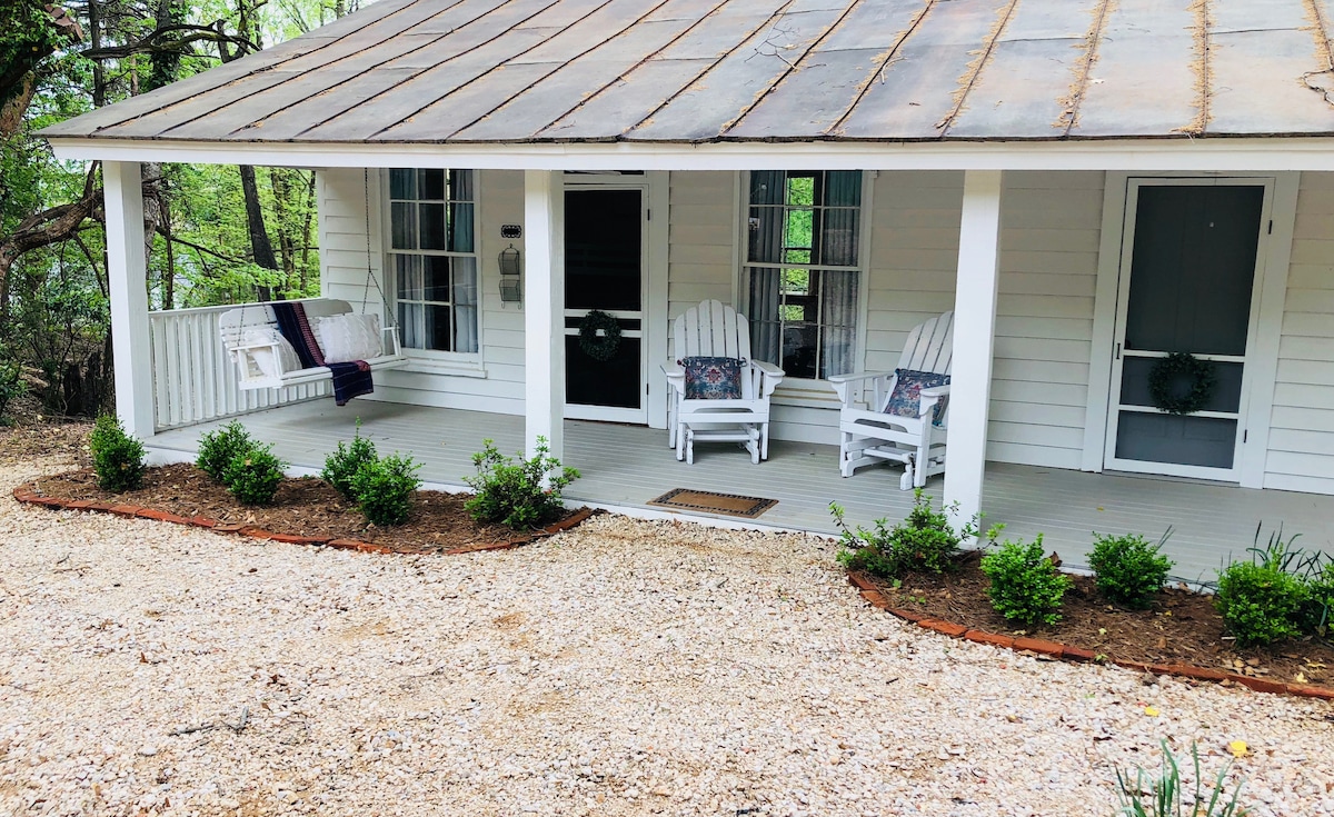 A welcoming front porch features two white rocking chairs and decorative pillows, surrounded by landscaped gravel pathways and low shrubs. The wooden structure displays a rustic metal roof and large windows, allowing natural light to illuminate the space.
