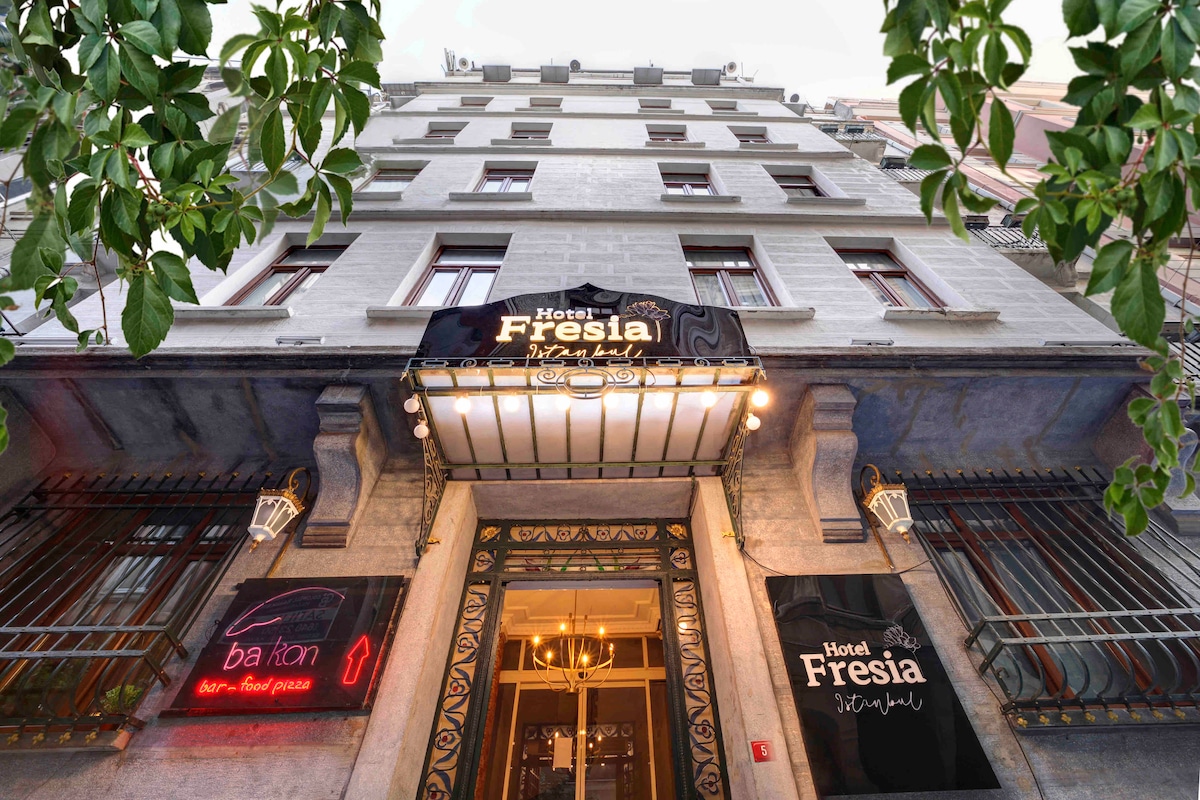 The entrance of Hotel Fresia is highlighted by a distinct awning displaying its name, framed by elegant architectural details. Potted plants can be seen nearby, adding a touch of greenery to the entrance. The façade features large windows and decorative elements that enhance the building's character.