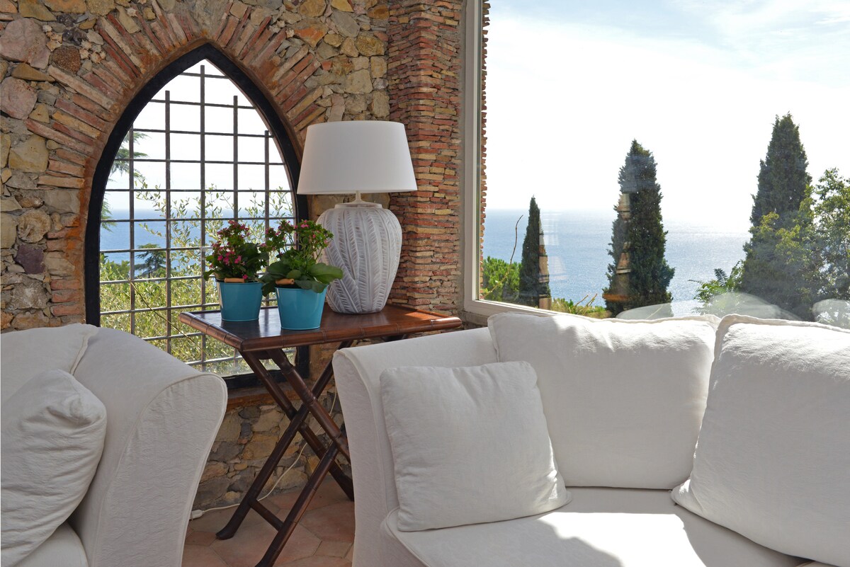 A cozy seating area features a light-colored sofa alongside a wooden table with potted plants. A decorative lamp stands on the table. Large windows provide panoramic views of the sea and surrounding greenery.