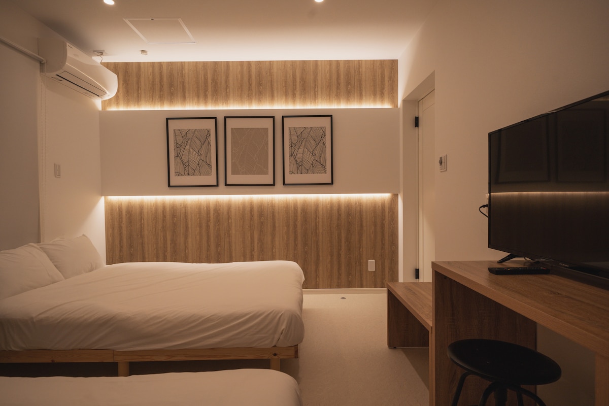 The room features two spacious beds with crisp white linens positioned next to each other. Soft ambient lighting highlights the wooden wall, complemented by framed artwork. A flat-screen TV and a streamlined desk are visible, contributing to the modern aesthetic of the space.