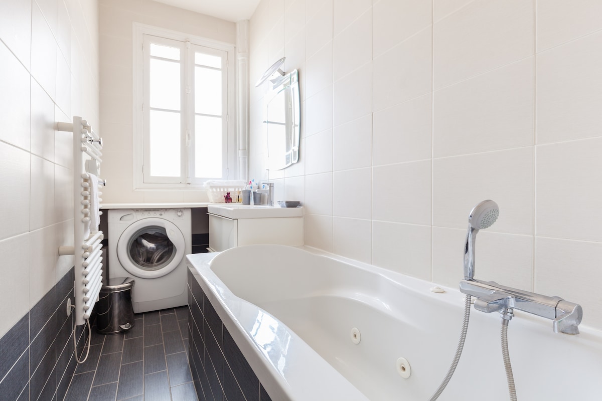 A modern bathroom is highlighted by a spacious bathtub and a washing machine. Natural light fills the room through a window. A sleek mirror and a sink are positioned above a white vanity. A towel radiator and a waste bin add functional elements to the space.
