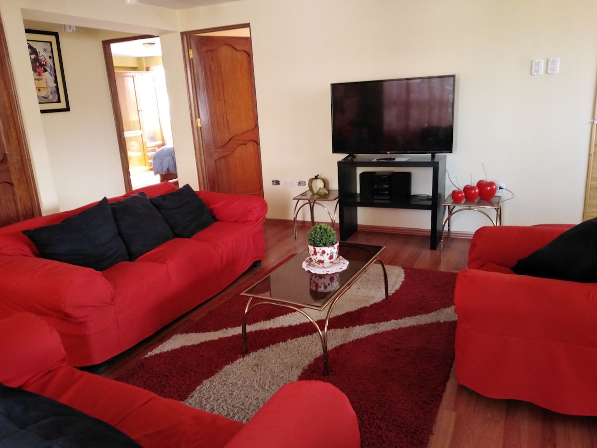 A living area features two red sofas with black cushions, arranged around a glass-top coffee table on a patterned rug. A television stands on a media console, and soft lighting contributes to a relaxed atmosphere.