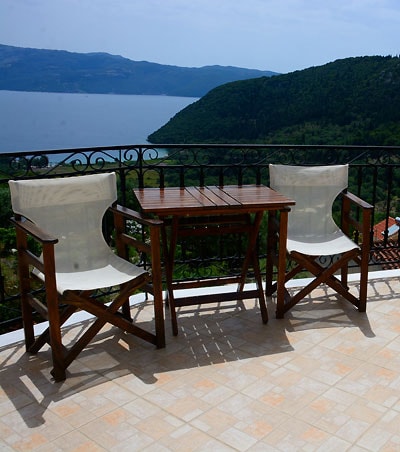 A small outdoor dining area features a wooden table accompanied by two folding chairs. The space is enclosed by a wrought iron railing, providing an unobstructed view of the sea and surrounding hills. Natural light highlights the textures of the surface beneath.