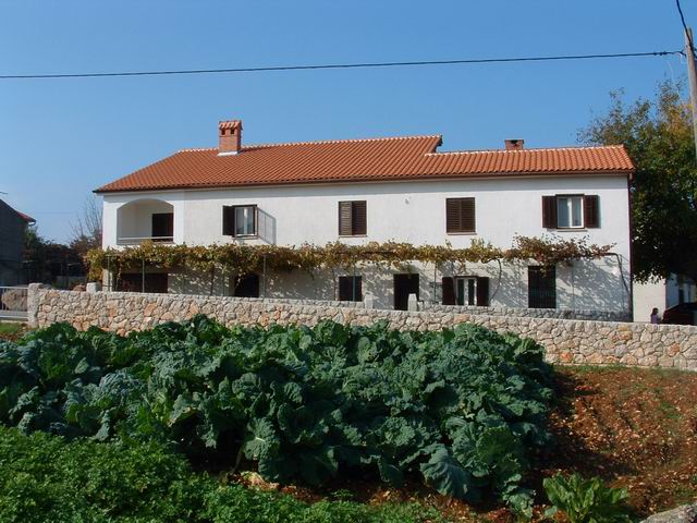 A two-story building is framed by a stone wall, featuring multiple windows and a red-tiled roof. Beneath a clear blue sky, a vegetable garden with leafy green crops is cultivated in the foreground, creating a natural contrast with the structure.