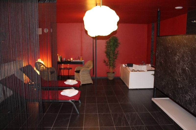 A wellness room features two lounge chairs positioned near a softly lit area, complemented by a small table with refreshments. A potted plant adds a touch of greenery, while a jacuzzi is visible in the background, surrounded by sleek black tiles and a modern partition.