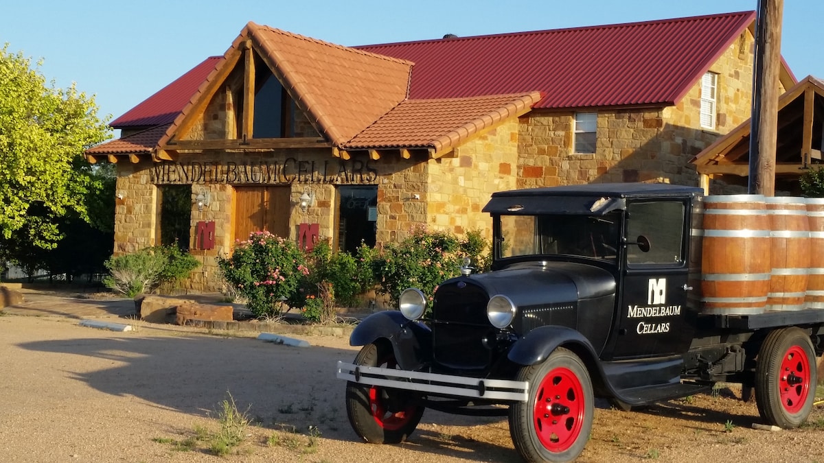 The entrance of Mendelbaum Cellars features a rustic stone facade and wooden accents under a red metal roof. A vintage truck with a wooden barrel parked in front adds charm, while surrounding greenery and colorful flowers create a welcoming setting.