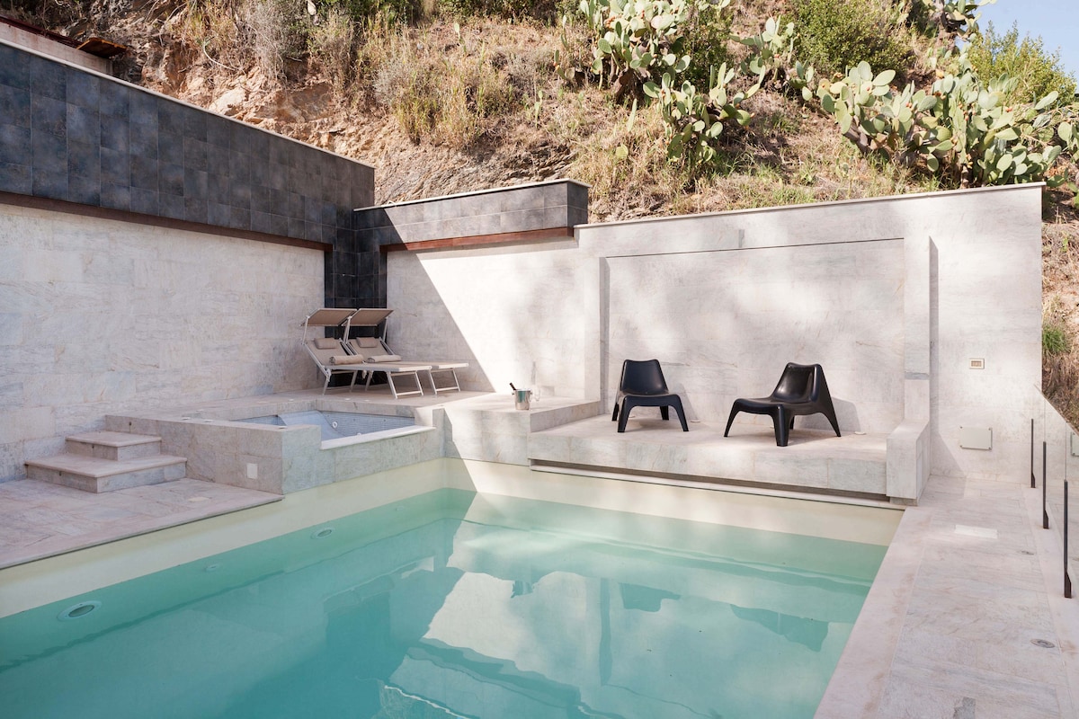 A private pool area features a well-maintained swimming pool with a set of modern black loungers positioned nearby. A jacuzzi sits adjacent to the pool, all surrounded by stone walls and natural landscaping, offering a serene space for relaxation.