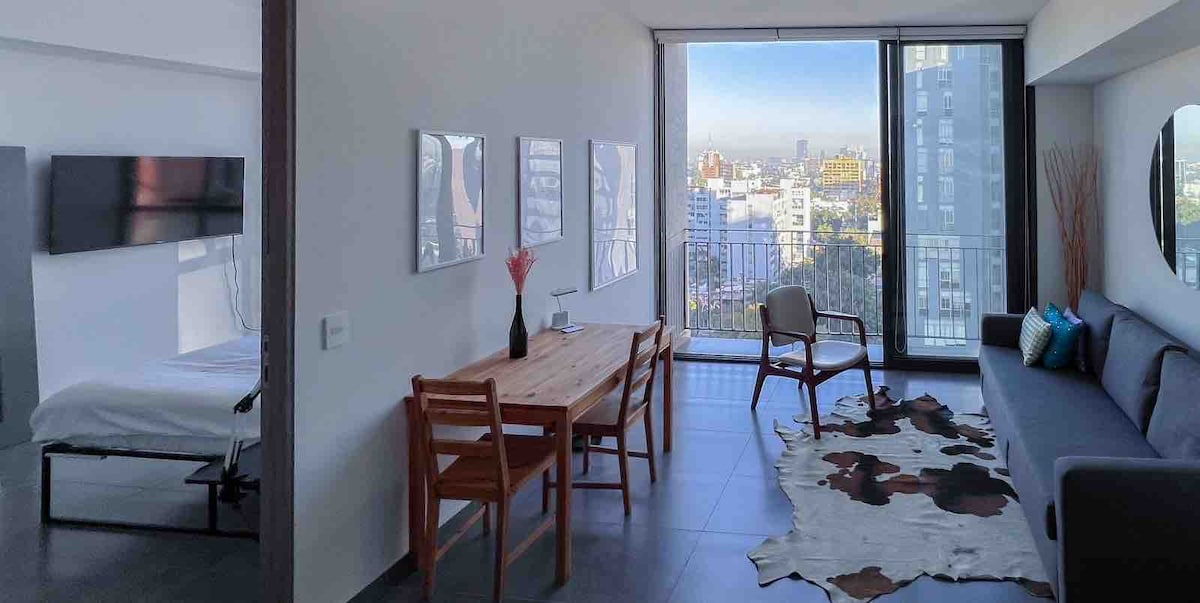 A spacious living area features a modern dining table with four wooden chairs. A comfortable sofa is positioned against the wall, while large windows provide a sweeping view of the city. Subtle decor enhances the minimalist aesthetic of the room.