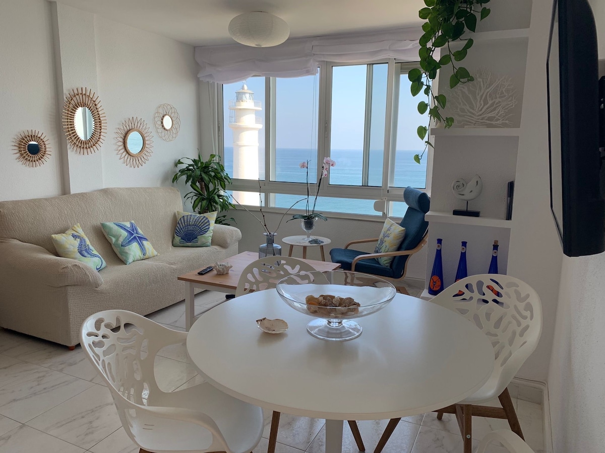 A bright living area features a comfortable sofa, overlooking the sea through large windows. A round dining table with white chairs sits in the center, complemented by wall mirrors and decorative accents. A potted plant adds a touch of greenery to the space.