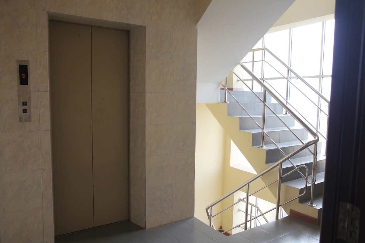 An elevator is positioned adjacent to a stairwell, both accessed from a spacious landing. The elevator features closed doors, while the stairs are enclosed by a metal railing and framed by large windows, allowing natural light to illuminate the area.