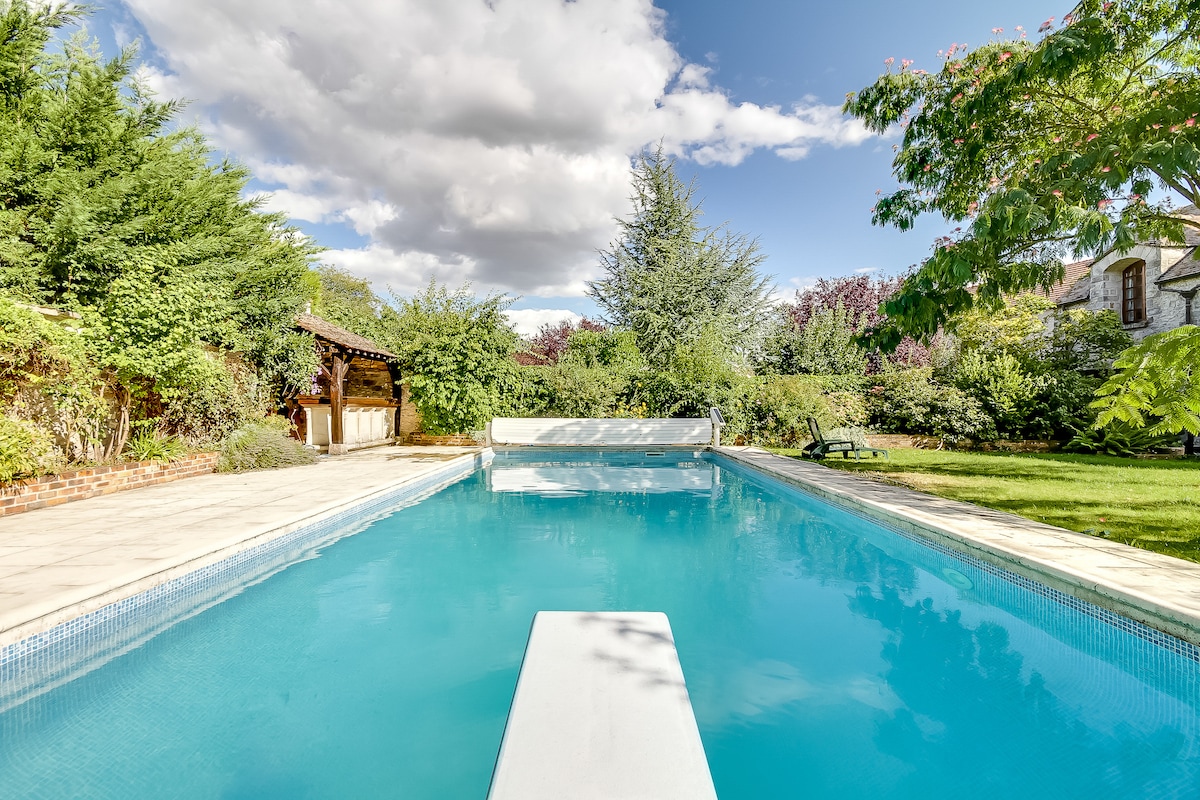 A serene outdoor scene features a large, non-heated swimming pool surrounded by lush greenery. The clear blue water reflects the sky, while vibrant trees and a garden add to the tranquil atmosphere. A shaded patio area with a wooden structure is visible in the background.