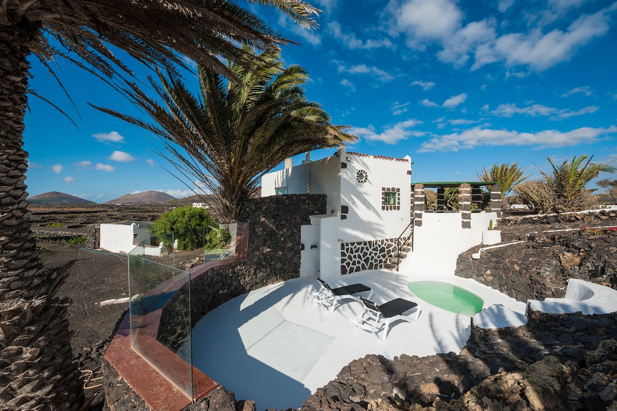 A small eco-house is set against a backdrop of volcanic landscape, featuring white walls and a private swimming pool. Large palm trees frame the area, while outdoor lounge chairs are positioned on a sun-soaked terrace, providing a serene space for relaxation.