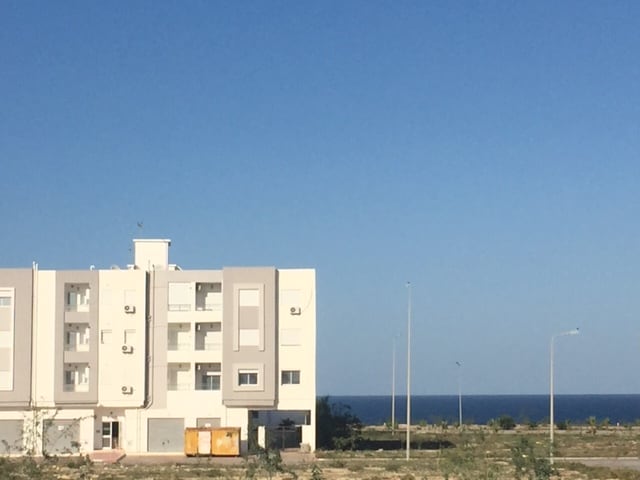 A newly constructed building is observed, showcasing a modern architectural design. Large windows reflect the clear blue sky, while the ocean is visible in the background. The surrounding area remains open, featuring minimal vegetation and few utility poles.