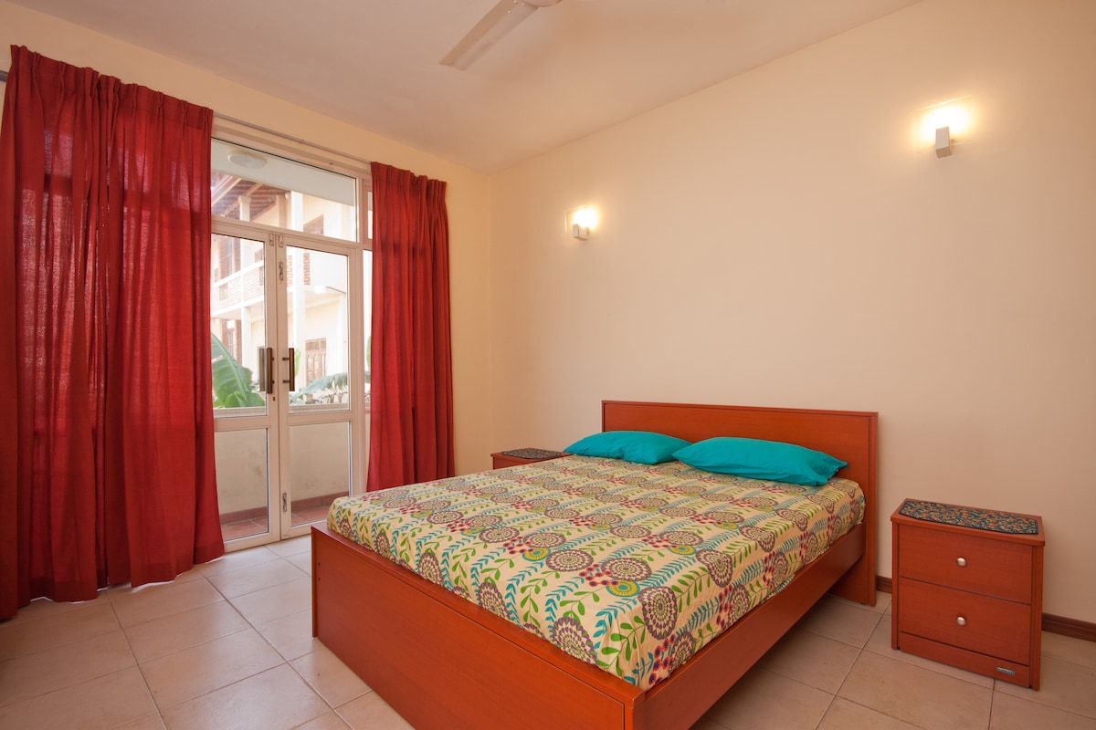 A well-lit bedroom features a spacious bed with a colorful patterned bedspread. Two blue cushions are placed on the bed. Large window views are framed by flowing red curtains, and a nightstand is positioned beside the bed.