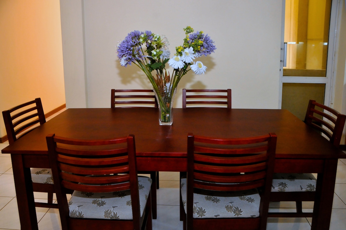 A wooden dining table is surrounded by six matching chairs, offering a spacious area for meals. A vase filled with colorful flowers creates a focal point in the center, complementing the neutral wall backdrop. Light from a nearby door enhances the setting.