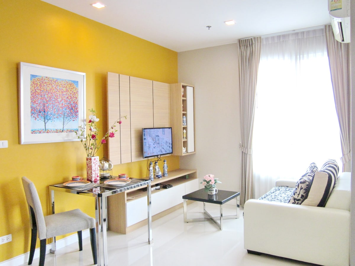 A modern living area features a white sofa and a glass coffee table positioned near a television. A stylish table is set with decorative items. Bright natural light floods the space through large windows, highlighting the warm yellow accent wall.