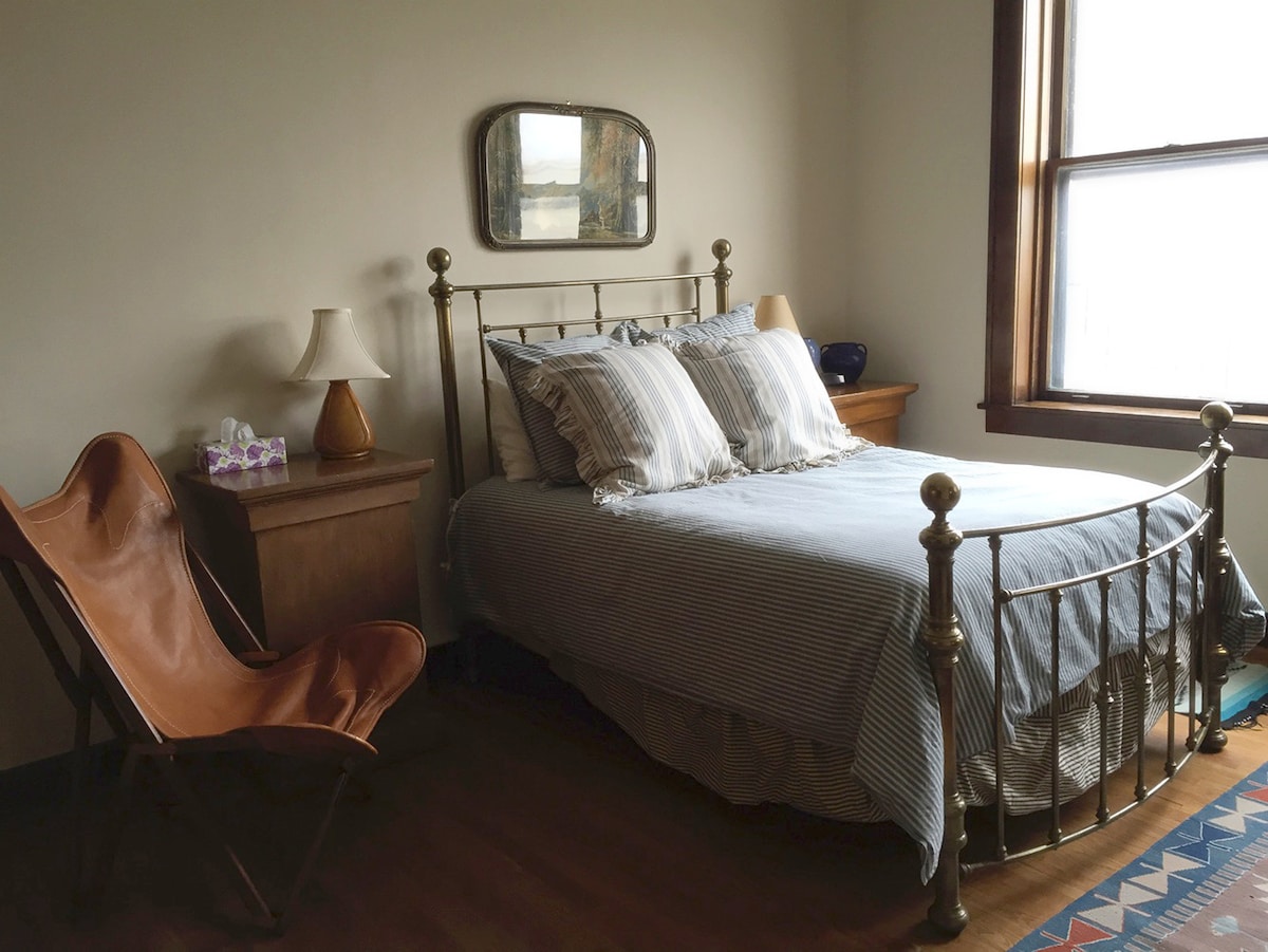 A bedroom displays a full-sized bed with navy and white striped bedding, flanked by two wooden nightstands. A vintage mirror hangs above the bed. A leather folding chair is positioned nearby, with a window allowing natural light to enter the space.