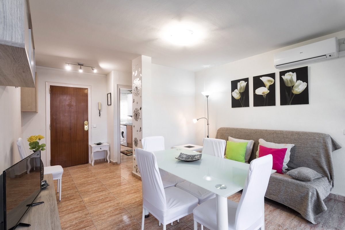 The living area features a soft gray sofa complemented by colorful throw pillows. A glass dining table with white chairs is positioned nearby, while a wall-mounted television and a floor lamp provide additional comfort. Light-colored decor enhances the bright atmosphere.