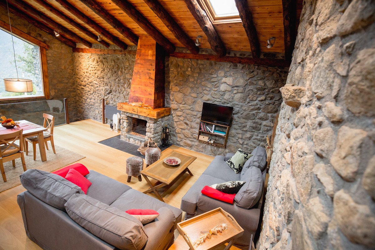 A spacious living area features stone walls and wooden beams, with a central fireplace adding warmth. Comfortably arranged are a gray sofa and armchairs, complemented by a wooden coffee table. A dining table is set nearby, illuminated by natural light streaming through a skylight.