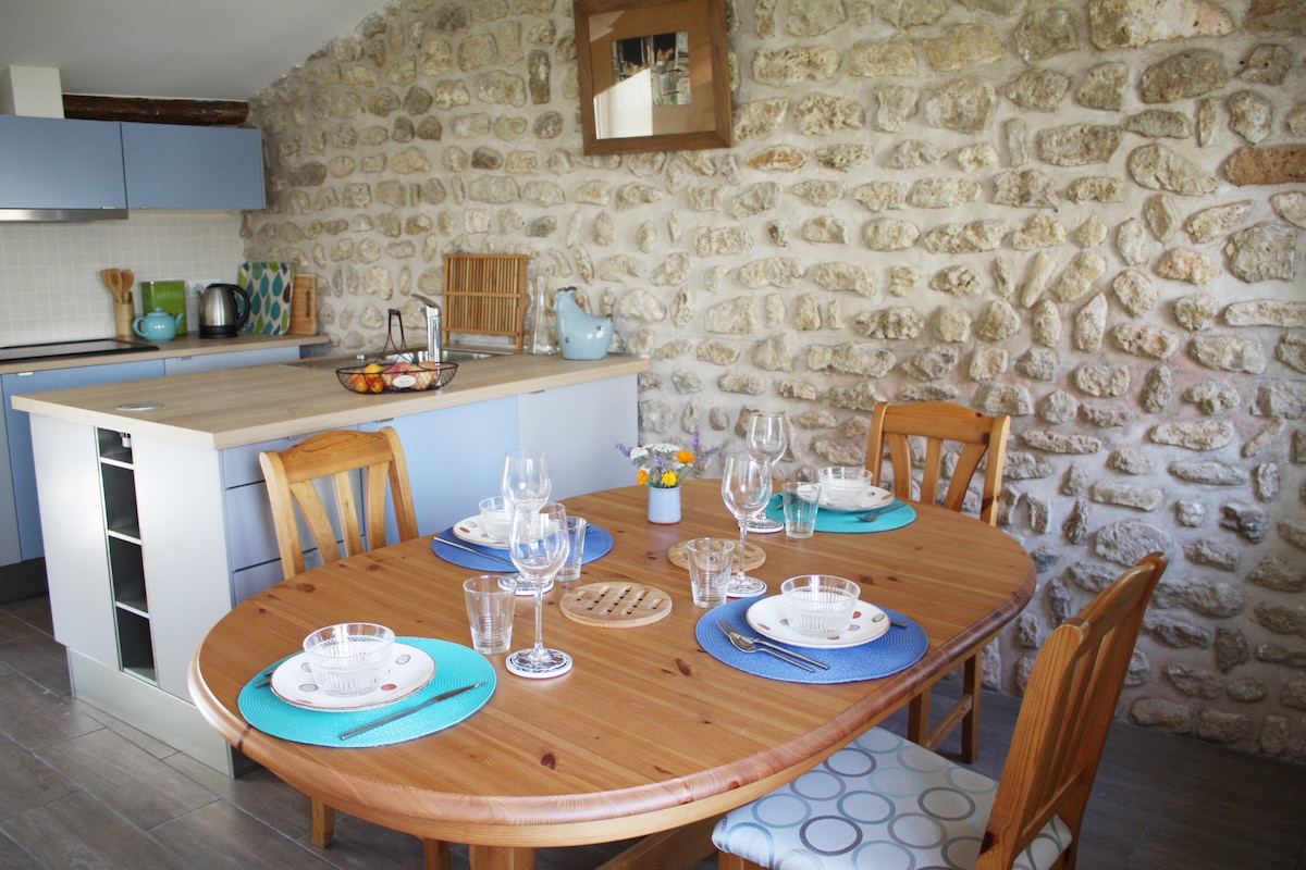 A wooden dining table is set with plates and glassware, surrounded by chairs. The room features a stone wall and a modern kitchen area with a light blue design. Natural light illuminates the space, enhancing the inviting dining atmosphere.