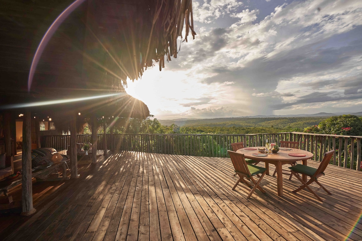 A spacious wooden deck offers panoramic views of the surrounding landscape, with the sun setting on the horizon. A circular table is set with chairs, inviting guests to enjoy meals outdoors in a serene environment.
