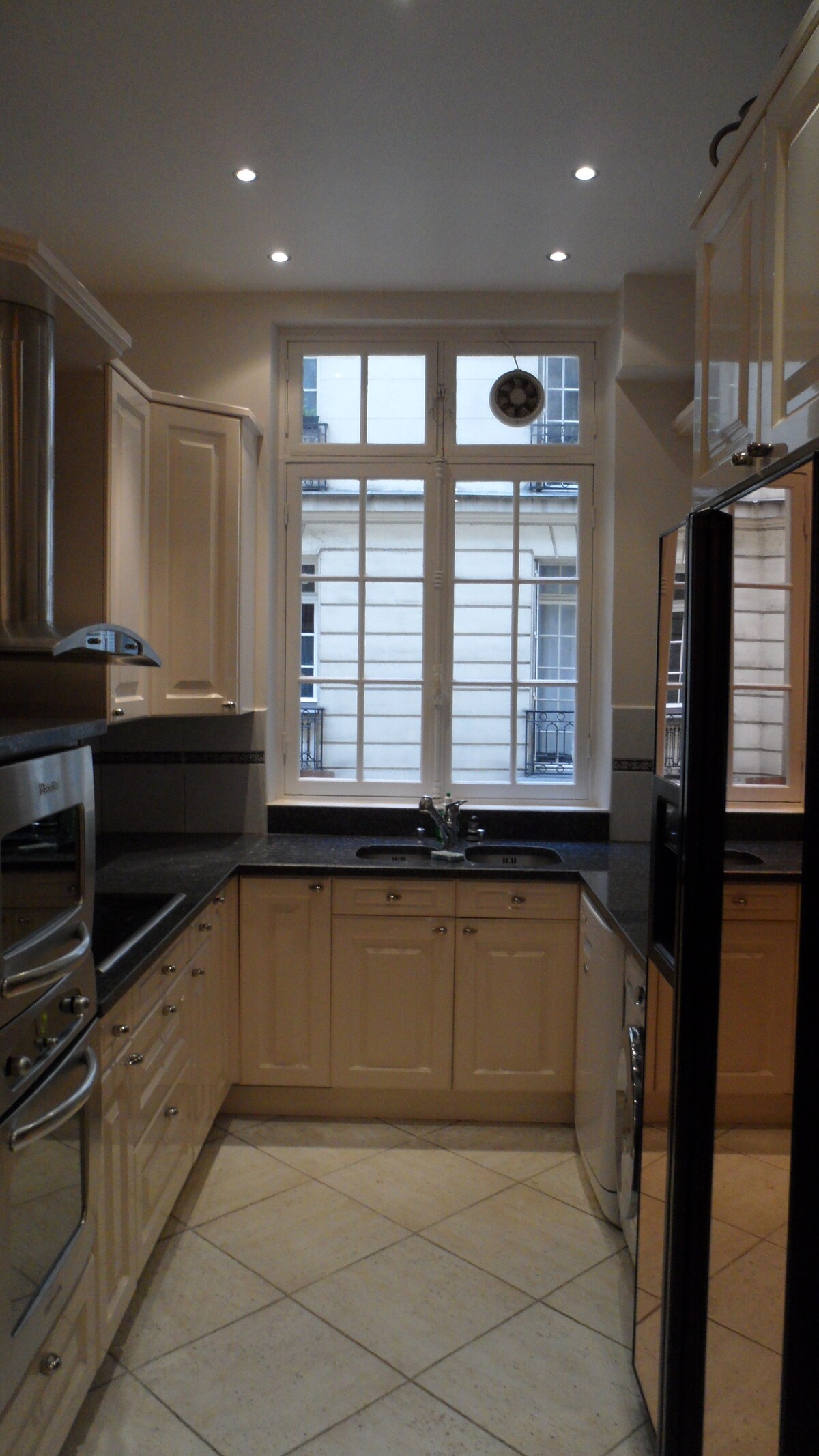 The kitchen features modern cabinetry with a sleek dark countertop. Light is provided by overhead fixtures and a large window, which looks out into the courtyard. Stainless steel appliances add a contemporary touch, alongside a washing machine discreetly integrated into the design.