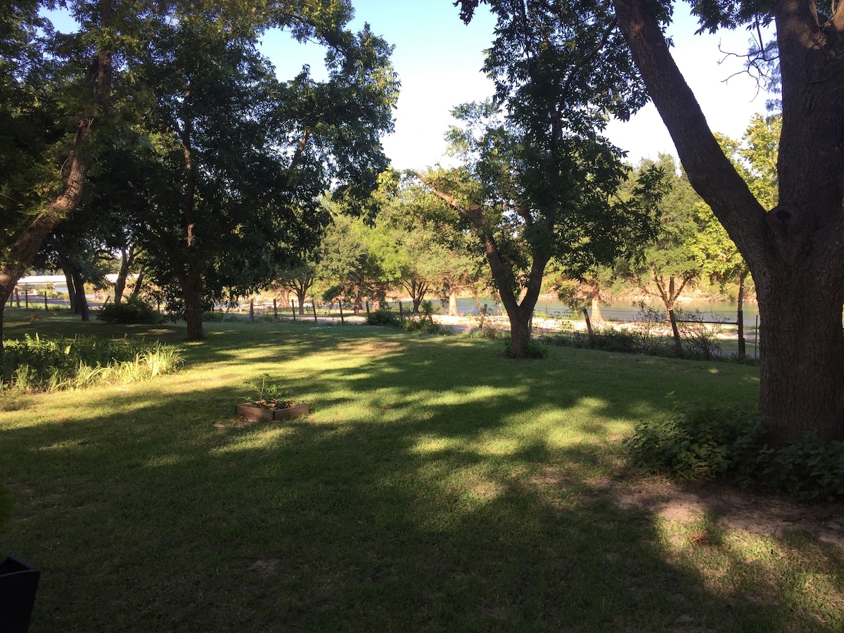 A spacious yard is covered in lush green grass and shaded by large pecan trees. The view extends towards the Blanco River, which is partially visible through the trees. Dappled sunlight creates a serene atmosphere across the landscape.