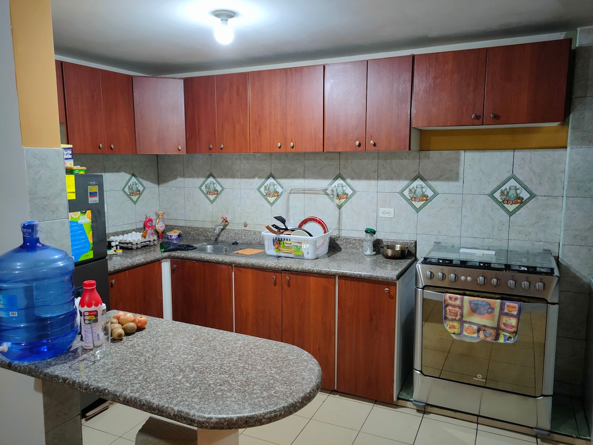 A modern kitchen features wooden cabinets and a spacious granite countertop. The area contains a gas stove, a stainless steel sink, and a variety of cooking essentials. Bright overhead lighting illuminates the room, which also includes a water dispenser and a collection of ingredients on the counter.