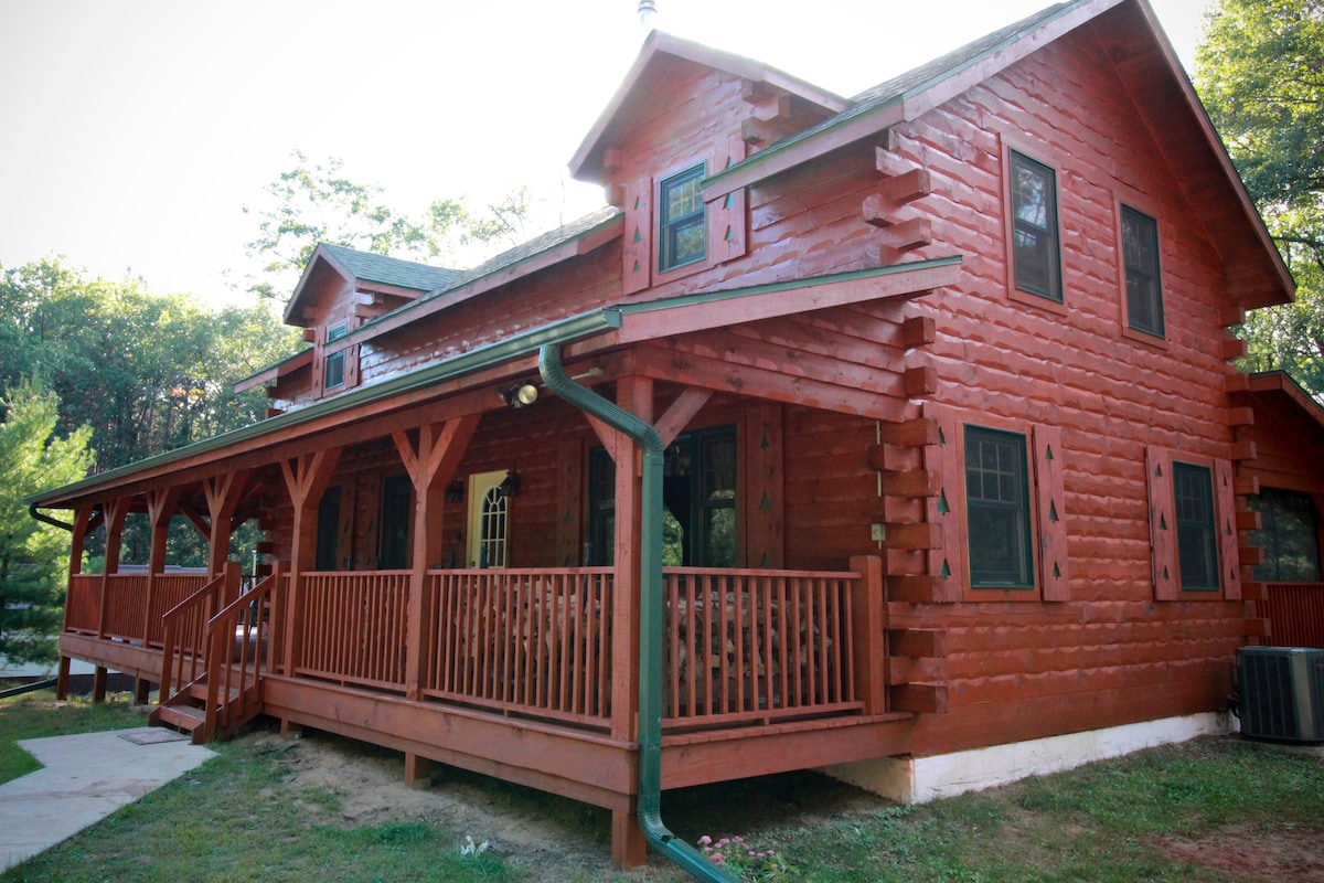 The exterior of a log cabin is displayed, featuring a rich red-brown wood finish. A spacious front porch with wooden railings wraps around the cabin. Large windows allow natural light to enter, while lush greenery surrounds the property, enhancing its secluded feel.