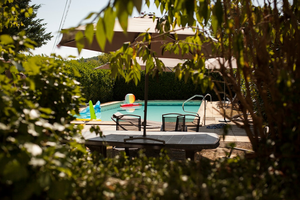 A view of the pool area is framed by lush greenery and umbrellas providing shade. Colorful pool floats rest on the water, while a stone table surrounded by chairs is visible, offering a place to relax and enjoy the surroundings.