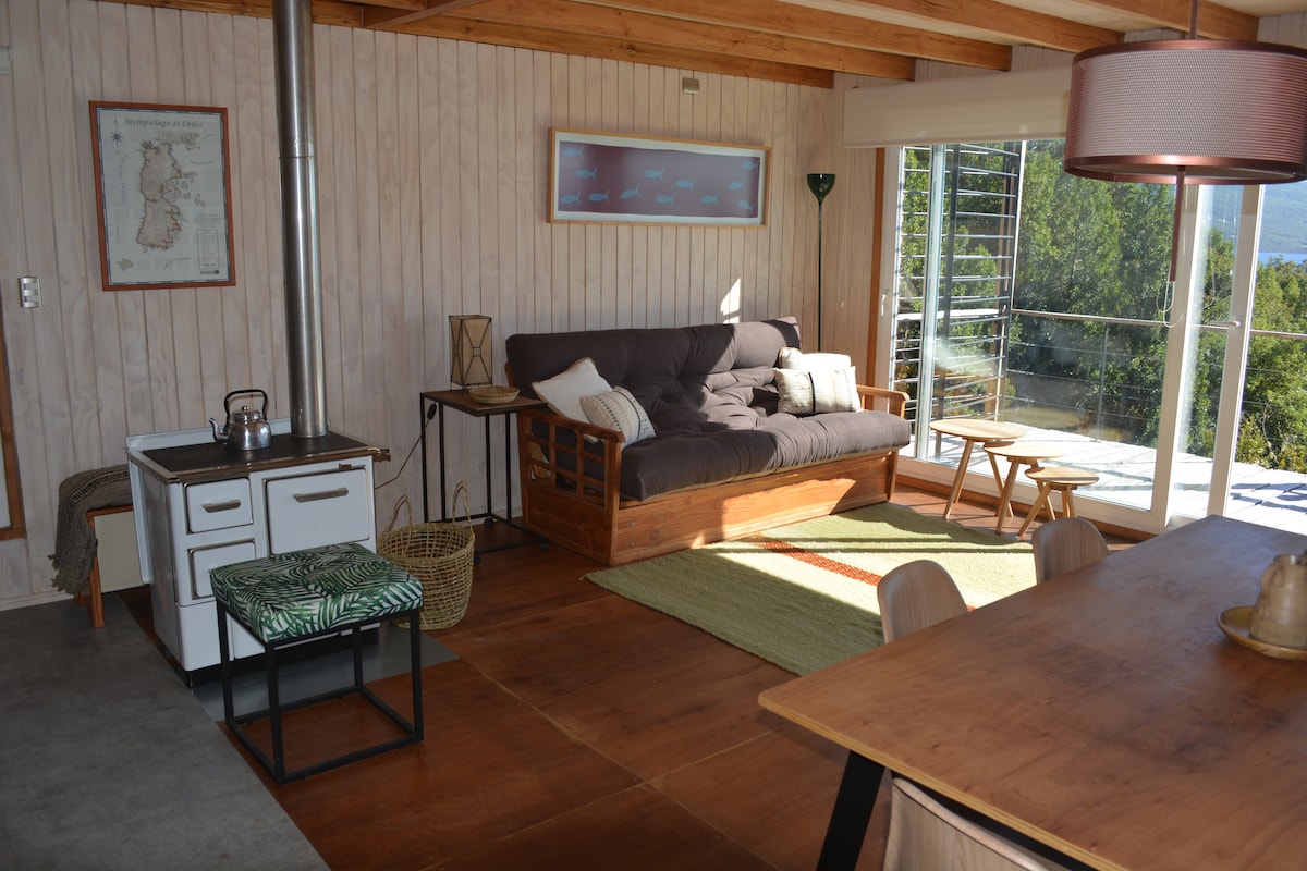 An inviting living area features a comfortable futon and a dining table. Natural light fills the space through large windows, with views of the surrounding greenery. A rustic wood-burning stove and a small kitchen area complement the cozy atmosphere.