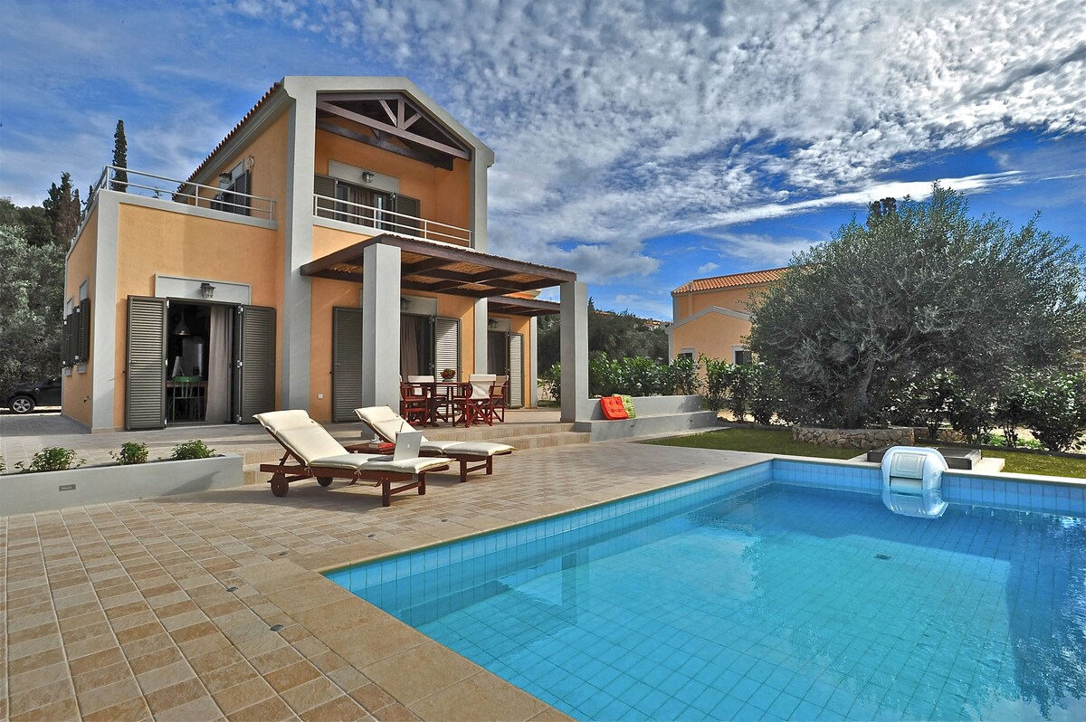 A two-storey villa is presented with a stone-tiled patio surrounding a private pool. Sun loungers invite relaxation, while a dining area with a wooden table is positioned under a shaded porch. Lush greenery and olive trees enhance the tranquil outdoor setting.