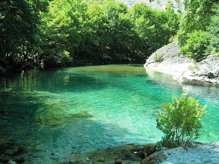 A serene natural pool is surrounded by lush greenery, with clear blue-green water reflecting the light. The shoreline is marked by smooth stones and small plants, creating a tranquil setting among the trees.