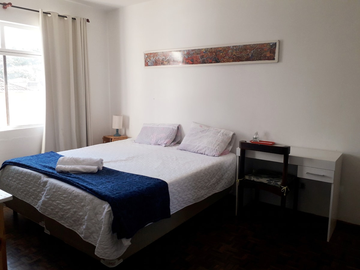 A bright sleeping area features a spacious bed dressed in crisp white linens. A blue throw is neatly placed at the foot of the bed. A bedside lamp and a small nightstand are positioned beside the bed, with a simple desk and chair nearby. Natural light enters through a window framed by light curtains.