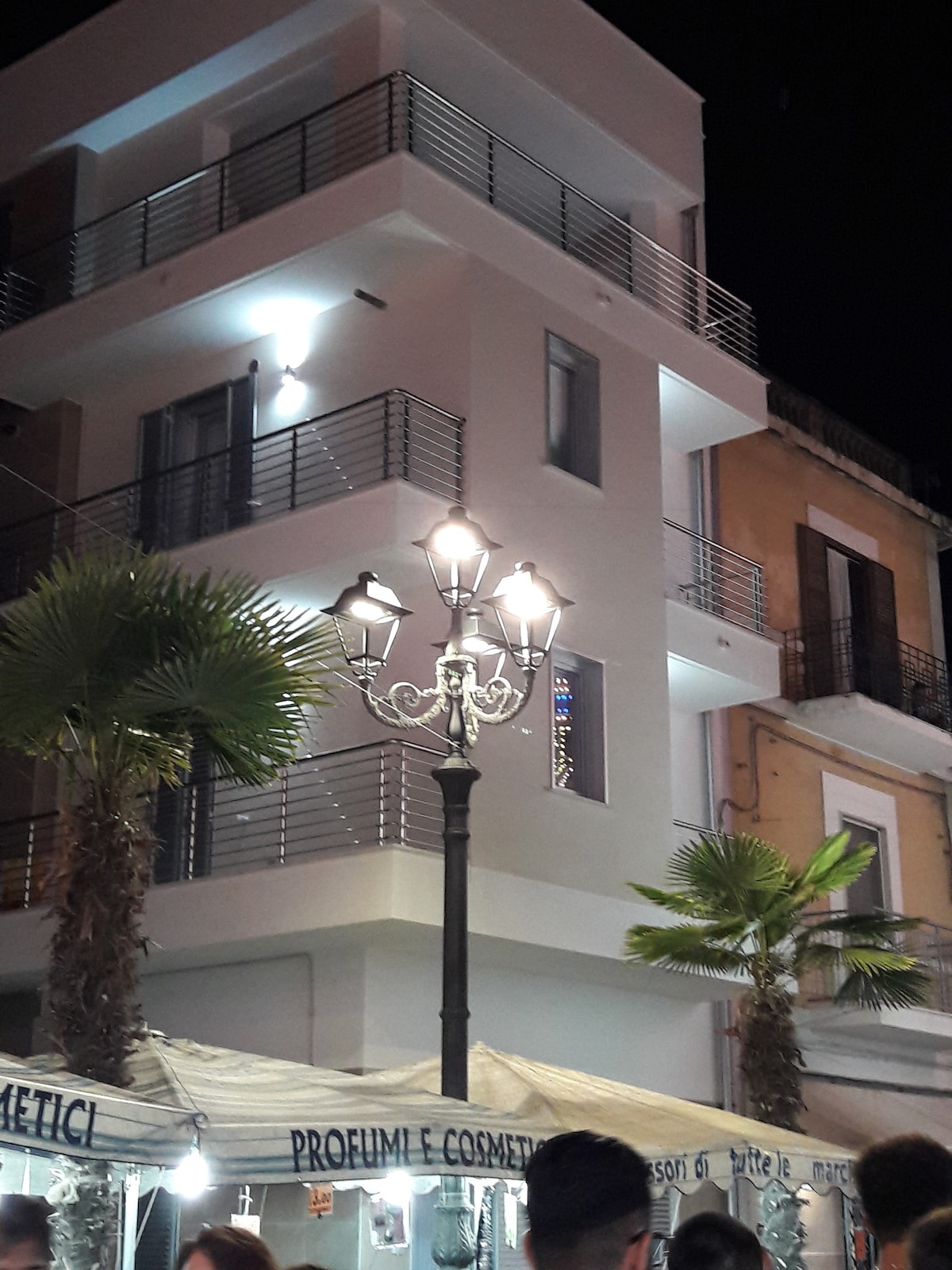 A modern three-story building features a sleek white façade with large windows and balconies. Decorative palm trees line the sidewalk, and a vintage street lamp adds charm. The scene is illuminated at night, showcasing the vibrant atmosphere of the surrounding area.