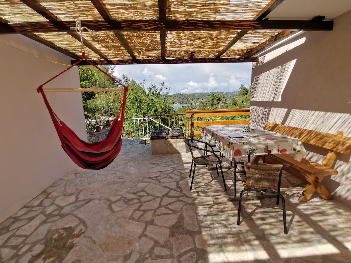 A spacious terrace features a large dining table and several chairs, inviting outdoor dining. A hammock is hung nearby, providing a relaxing spot. The area is shaded by a woven canopy, allowing soft natural light to filter through, with greenery visible in the background.