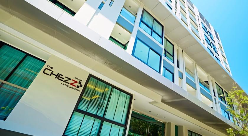 The exterior of The Chezz Metro Life Condominium is depicted, showing a modern building with large windows and a sleek design. Sunlight reflects off the glass, and the property name is prominently displayed on the wall, surrounded by lush greenery.