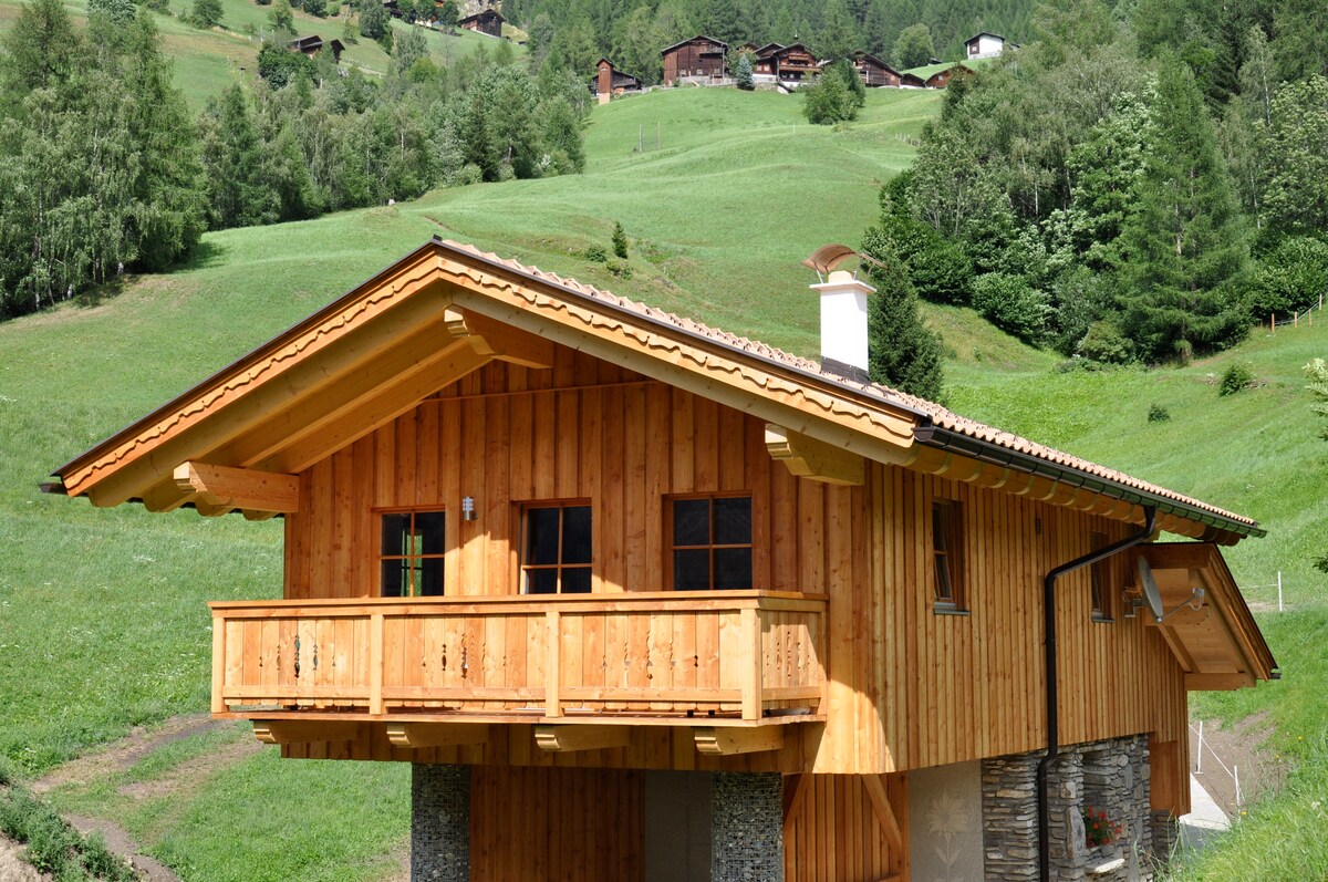 A wooden chalet features a sloped roof and a balcony, framed by lush green hills and forested areas. Natural stone accents complement the wooden structure, while large windows invite natural light into the interior.