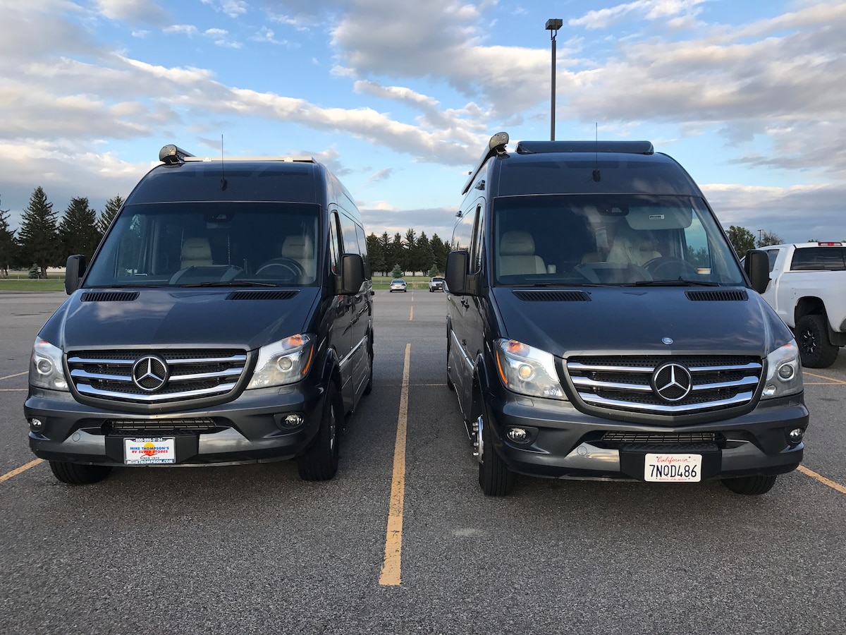 Two Mercedes Sprinter campervans are parked side by side in a lot. Both vehicles feature a sleek, modern design with tinted windows and roof racks. They are finished in a dark color scheme, with the front showing prominent grills and headlights.