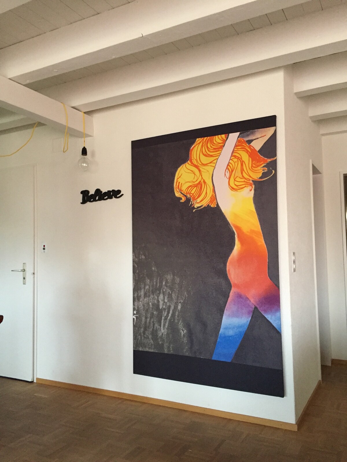 A vibrant mural of a figure with flowing hair and a gradient-colored body adorns the wall, adding a bold artistic touch to the living space. Subtle lighting fixtures and a door are visible nearby, contributing to an open and airy ambiance.