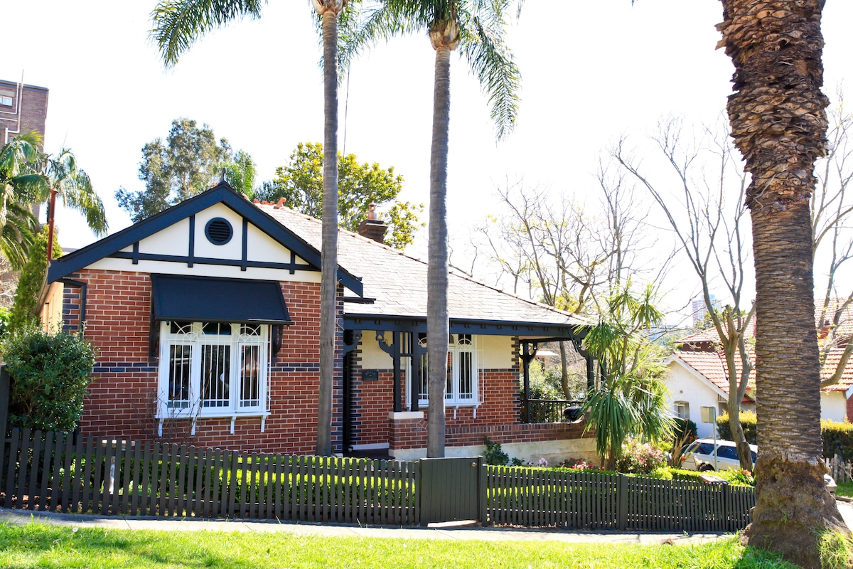 A charming heritage house is depicted with a red brick facade, white window frames, and a black awning. Lush palm trees frame the front yard, which is bordered by a black picket fence, enhancing the inviting exterior of the property.