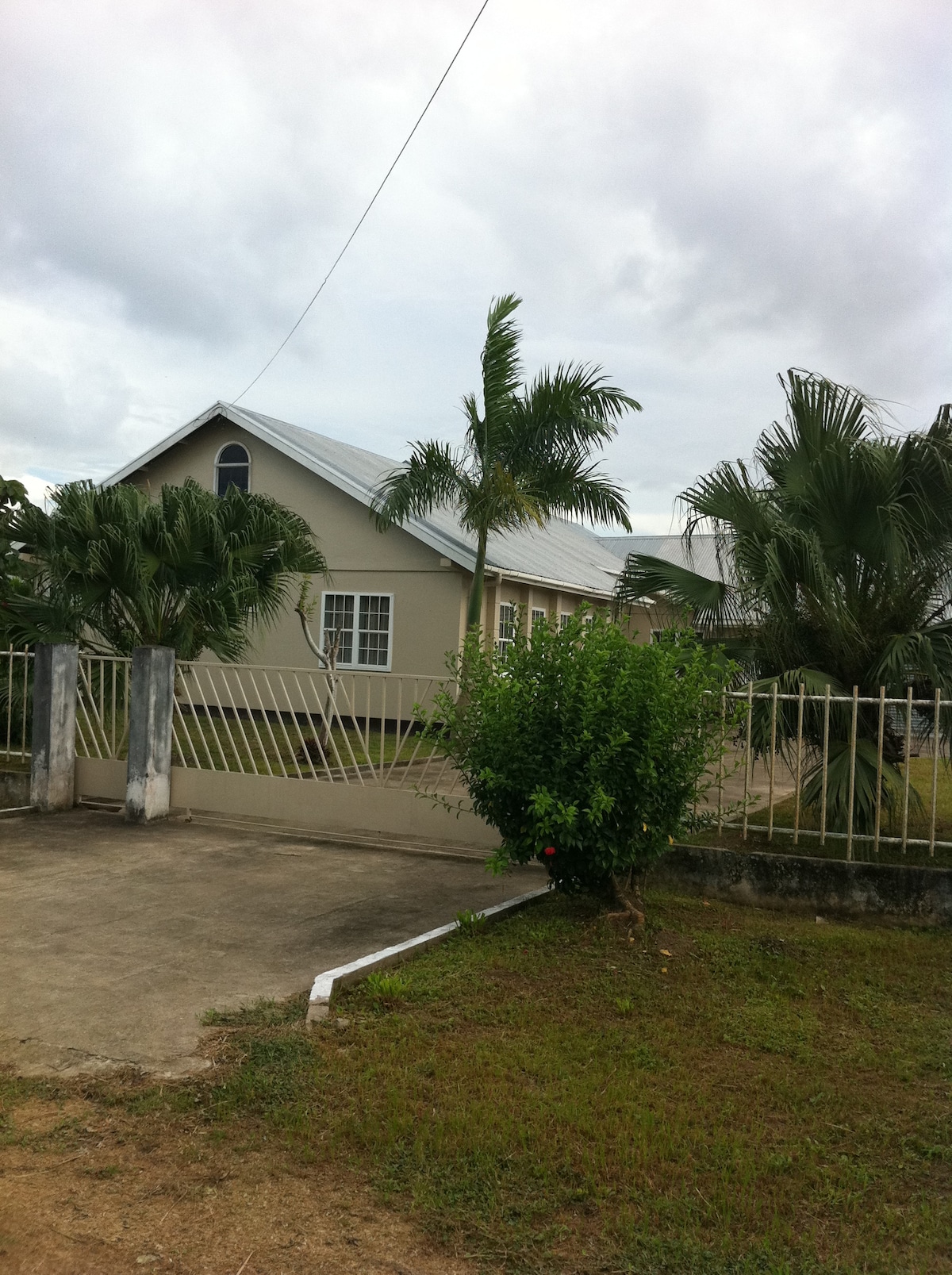 A modest three-bedroom villa is seen through a gated entryway, surrounded by greenery. The house features a light-colored exterior, a sloped roof, and multiple windows. Palm trees and other plants frame the property, adding to its serene setting.