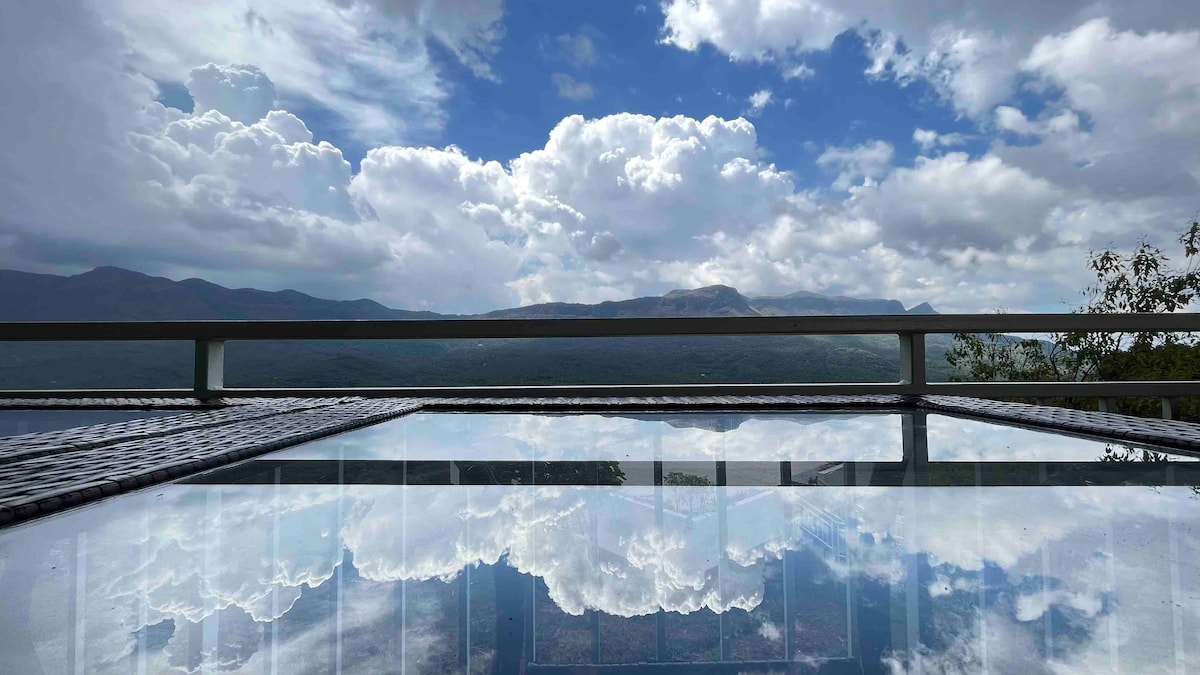 A serene view of the landscape is reflected in the calm surface of a pool. The backdrop features lush hills and a dramatic sky filled with fluffy clouds, creating a peaceful atmosphere. The balcony railing provides an unobstructed sight of the scenic surroundings.