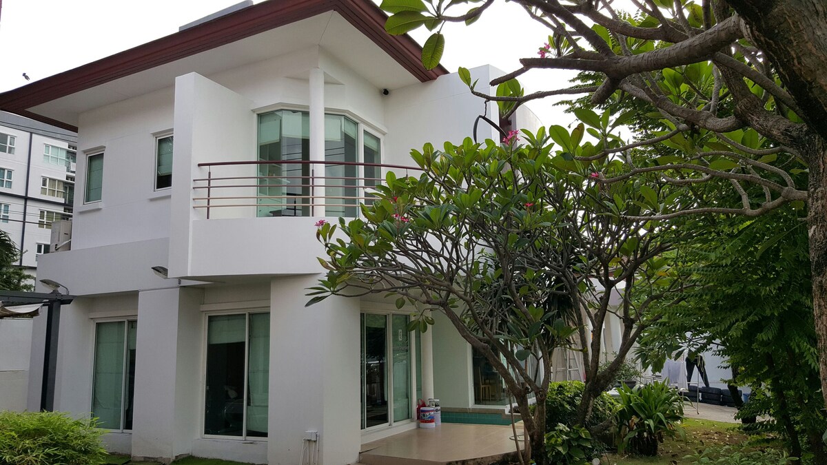 The exterior of a contemporary white house is shown, featuring large windows and a second-floor balcony. Lush greenery surrounds the structure, including a flowering tree and various shrubs, creating a serene environment. The entrance is accessible through a paved pathway.
