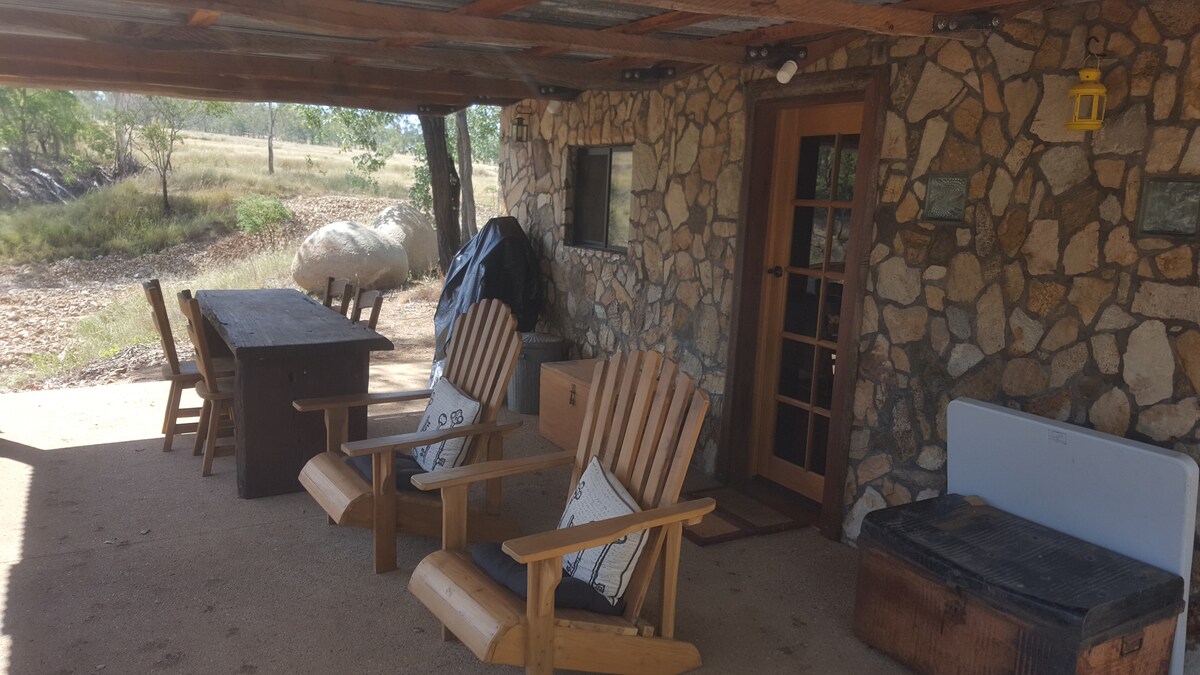 An outdoor seating area is presented, featuring two wooden chairs and a table made of rustic materials. A bench is positioned near a cooler. The stone wall provides a textured backdrop, while the surrounding landscape includes grassy areas and trees.